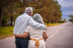 Couple d'aînés marchant enlacés de dos le long d'une route bordée d'arbres.