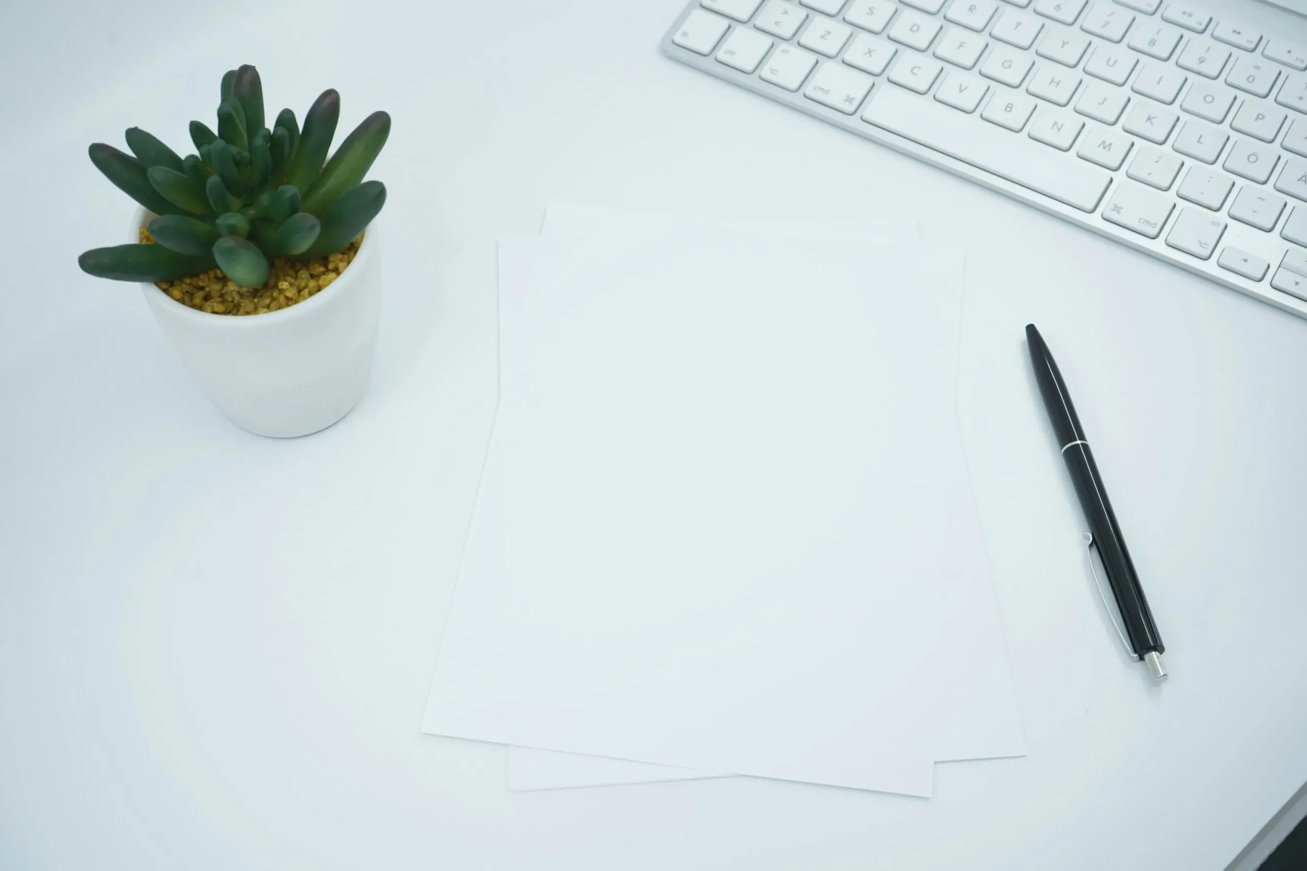 Bureau de travail blanc avec feuilles vierges, stylo, clavier et plante.